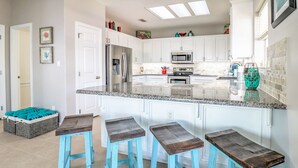 A kitchen with white cabinets, a granite countertop, and a stainless steel refrigerator. There are three blue stools and a basket of teal towels. - Southern Destination: Bask longer, save 10% on stays of 7+ nights! (South Padre Island)