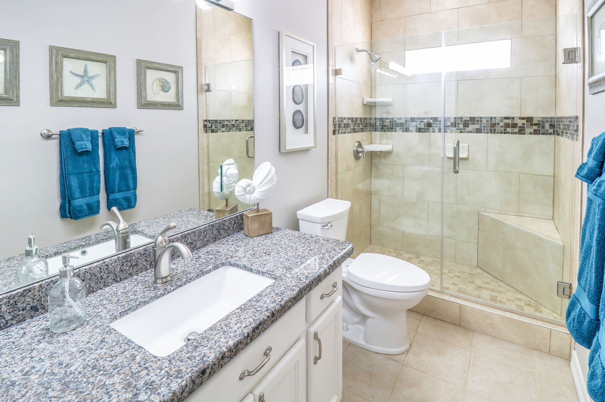 Un baño con lavamanos de encimera de granito, inodoro blanco, ducha con mampara de cristal y toallas azules colgadas de ganchos.