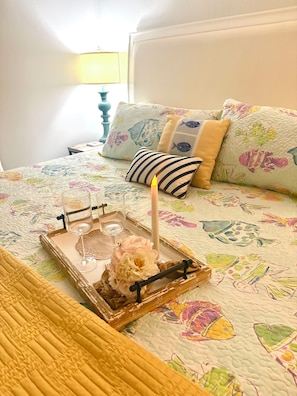Un lit recouvert d’une courtepointe à motifs floraux, une table de chevet avec une lampe et un plateau avec une bougie et des verres.