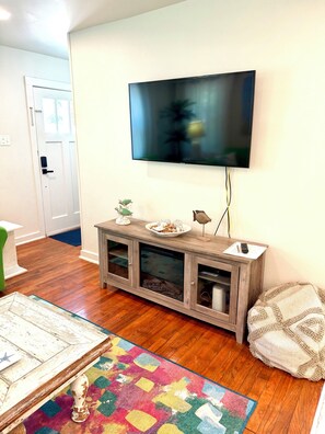 A living room with a flat-screen TV mounted on the wall, a wooden cabinet with decorative items, a colorful rug, and a door leading to another room.
