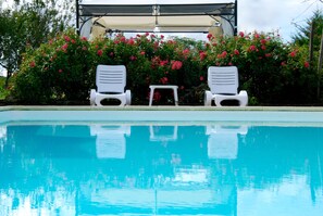 Piscina com duas cadeiras de praia brancas e uma pequena mesa sob um toldo, cercada por arbustos verdes exuberantes com flores cor-de-rosa.