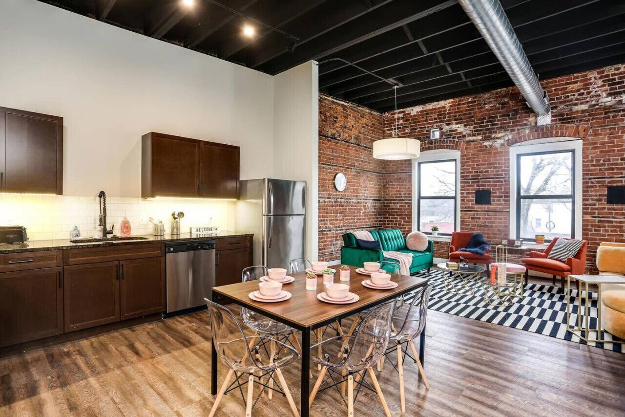 A modern kitchen with stainless steel appliances, a dining area with a checkered floor, and a seating area with a brick wall and large windows.