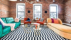 A room with a brick wall, two large windows, a green sofa, an orange sofa, and a black and white striped rug.