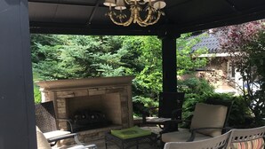 An outdoor patio with a stone fireplace, metal chairs, and a hanging light fixture.