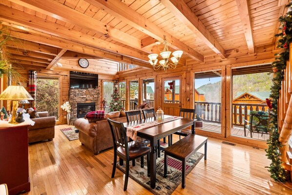 A cozy log cabin interior with a stone fireplace, wooden dining table, and a Christmas tree.
