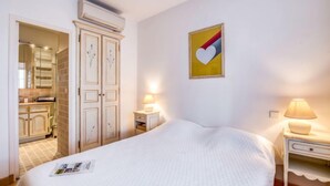 Une chambre à coucher comprenant un lit, des tables de chevet, une armoire et un coin cuisine visible à travers une porte ouverte.