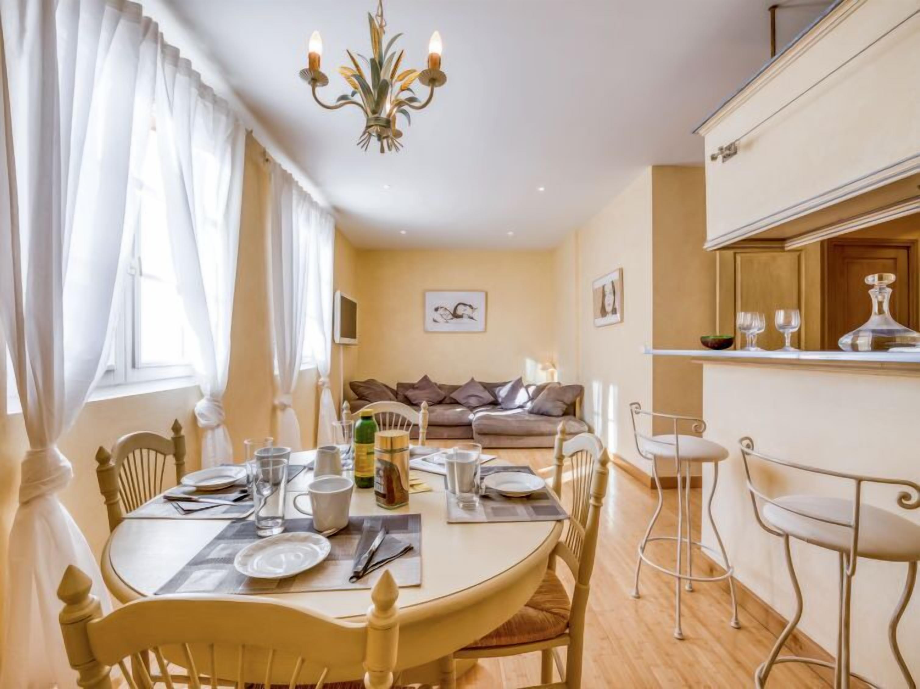 Un comedor con una mesa redonda dispuesta para cuatro personas, un sofá al fondo y una zona de bar con mostrador y taburetes.
