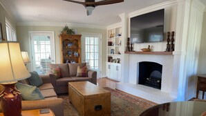 Un salon comprenant un canapé, une table basse, un pouf et une cheminée. Un téléviseur est fixé au-dessus de la cheminée, et un ventilateur de plafond est installé au plafond.