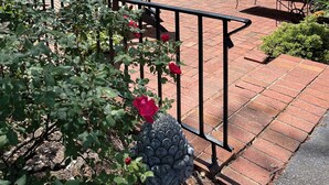Une terrasse extérieure avec un sol en briques, entourée d’une végétation luxuriante et d’arbres. On y trouve plusieurs tables et chaises, dont certaines sont dotées de parasols, et un bâtiment à l’arrière-plan, dont la façade est blanche.