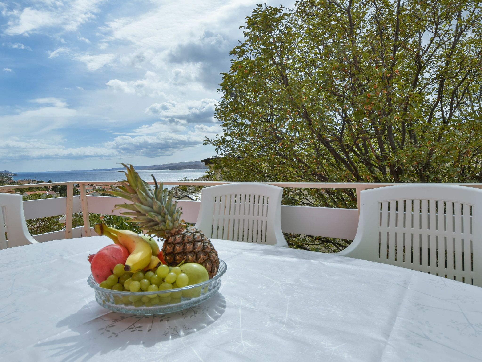 A table with a fruit bowl on a white tablecloth, overlooking a scenic view of the sea and a tree.