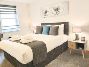A neatly made bed with white linens and a grey blanket, a bedside table with a lamp, and a wall-mounted artwork of flowers. - Inverness City Centre Apartment with Elevator (Inverness)
