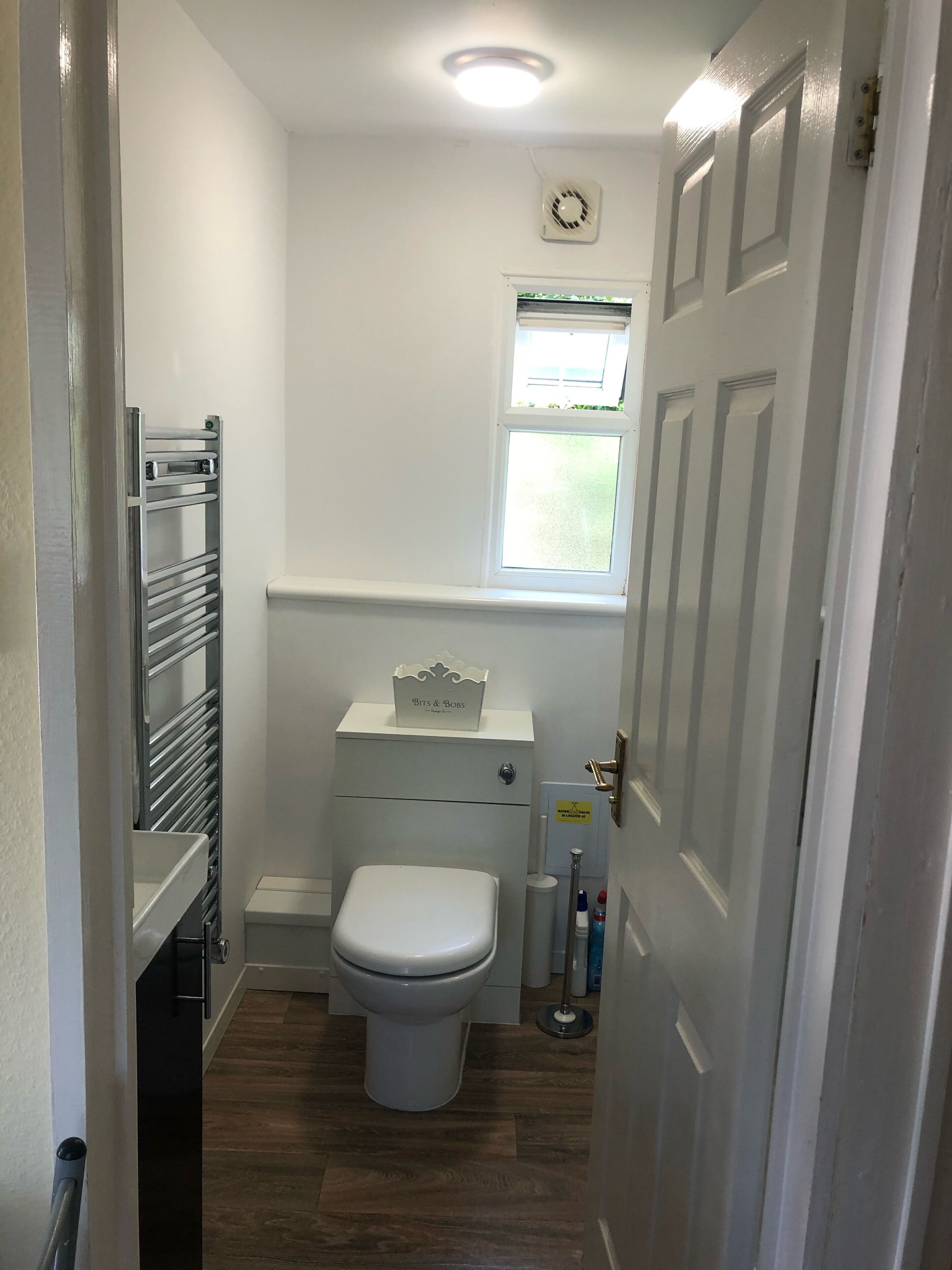 Un baño pequeño con inodoro, una ventana, un radiador y un estante con una caja etiquetada 'Bits & Bobs'.