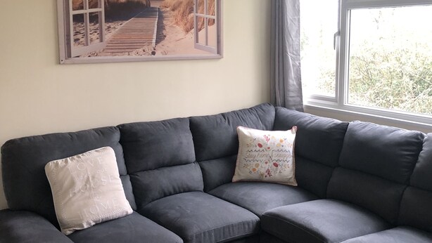 Ein graues Ecksofa mit zwei Zierkissen, ein Strandbild an der Wand und ein Fenster mit Vorhängen.