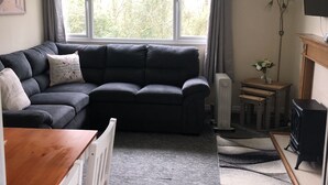 Ein Wohnzimmer mit einer dunkelgrauen Couch, einem Holzesstisch und einem weißen Stuhl. Es gibt einen Heizkörper, einen kleinen Beistelltisch mit einer Vase und eine schwarze TV-Konsole.