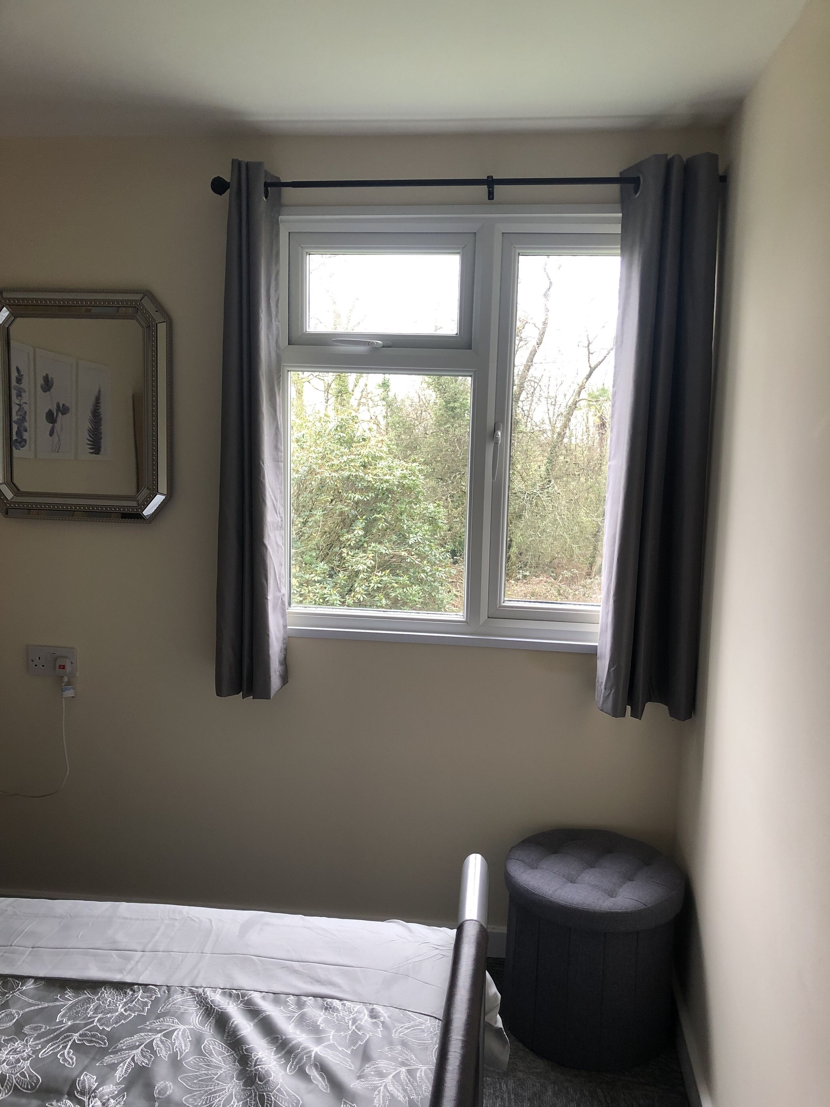 A bedroom with a bed, a window with curtains, a bedside table, and a wall-mounted power outlet.