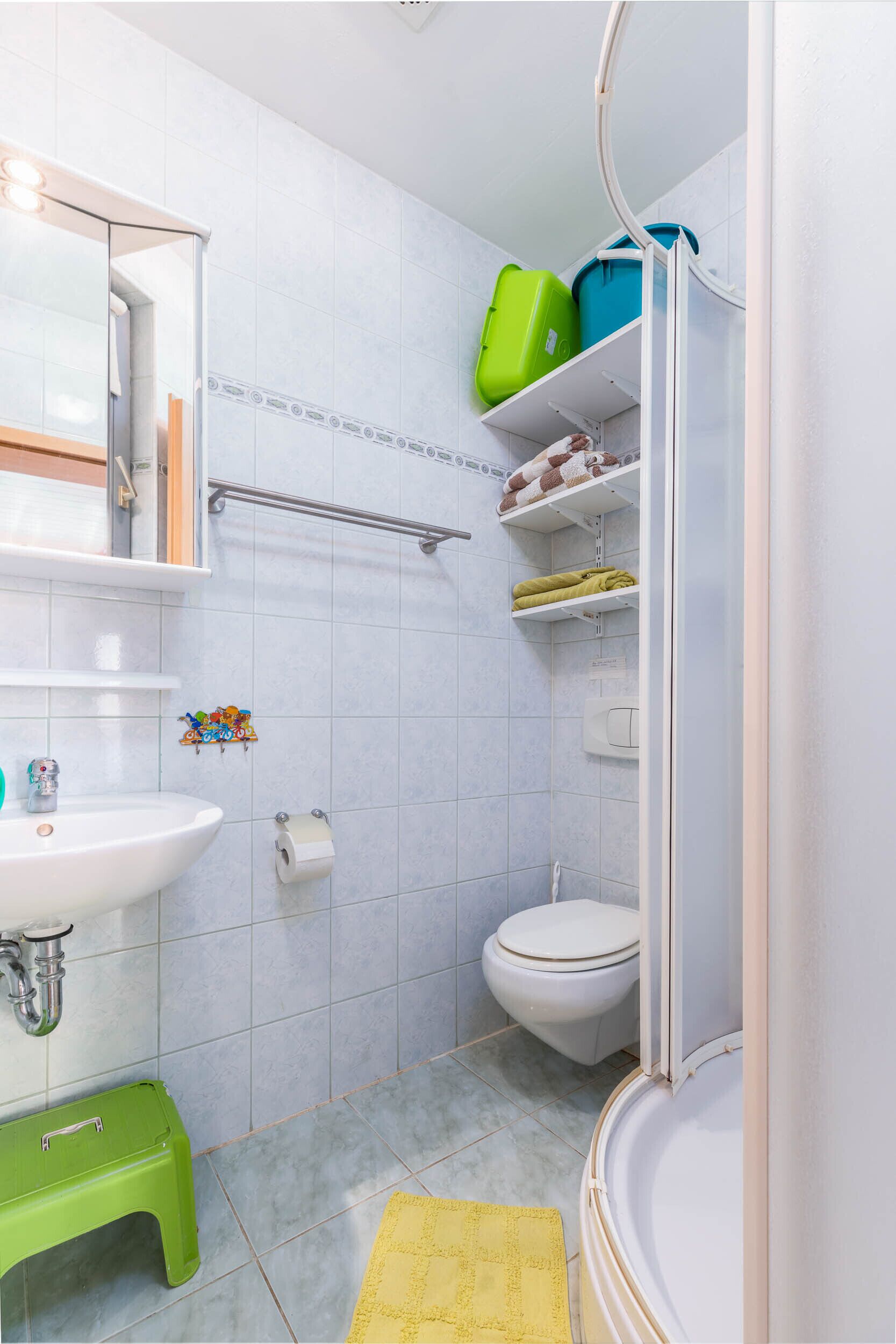 A bathroom with a sink, toilet, and shower area. There are shelves with toiletries and a green step stool.