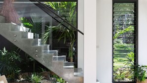 Un escalier moderne avec des rampes en verre, entouré de plantes d’intérieur et offrant une vue sur la verdure à travers une fenêtre.
