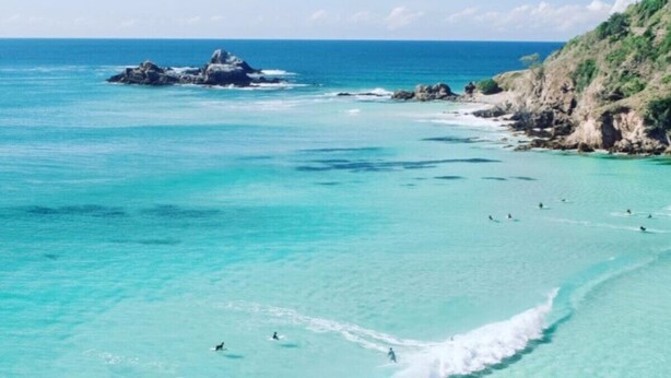 Une plage à l’eau turquoise et limpide, avec des rochers et quelques personnes qui se baignent et font de la planche à voile.