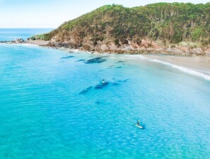 Vista costeira com águas turquesa cristalinas, uma praia arenosa e uma encosta arborizada.