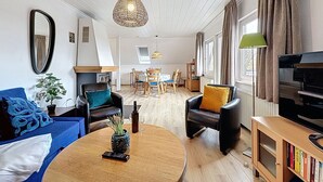 Un salon avec un canapé, des fauteuils, une table basse, un téléviseur et, au fond, un coin repas.