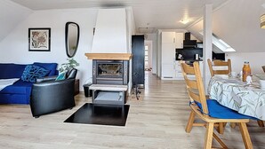 Un salon moderne avec une cheminée, un canapé, une table à manger avec des chaises, et un coin cuisine à l’arrière-plan.