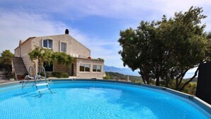 Una piscina con superficie de azulejos azul turquesa, una casa de fachada de piedra y vista al mar y colinas al fondo.