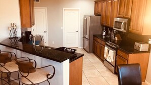 A kitchen with wooden cabinets, a black countertop, a refrigerator, a microwave, a coffee maker, and a toaster. There are three bar stools and a dining table.