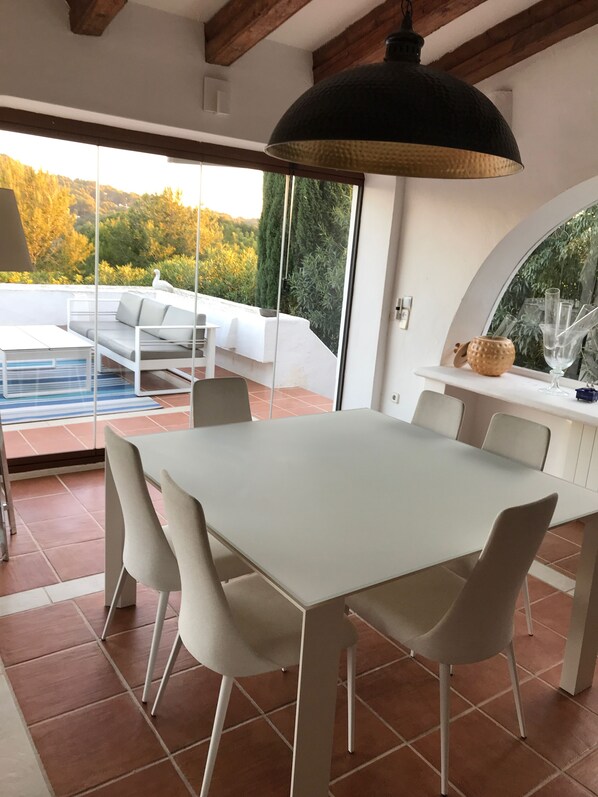 A modern dining area with a white table and chairs, a black pendant light, and a view of a garden through large glass doors.
