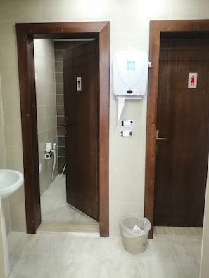 Casa de banho pública com secador de mãos, dispensador de papel higiénico e um caixote de lixo.