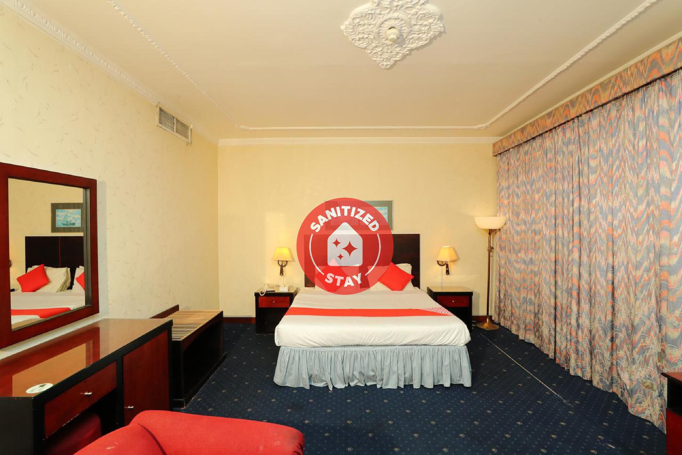 A hotel room with a large bed, a desk, a mirror, and a red chair.