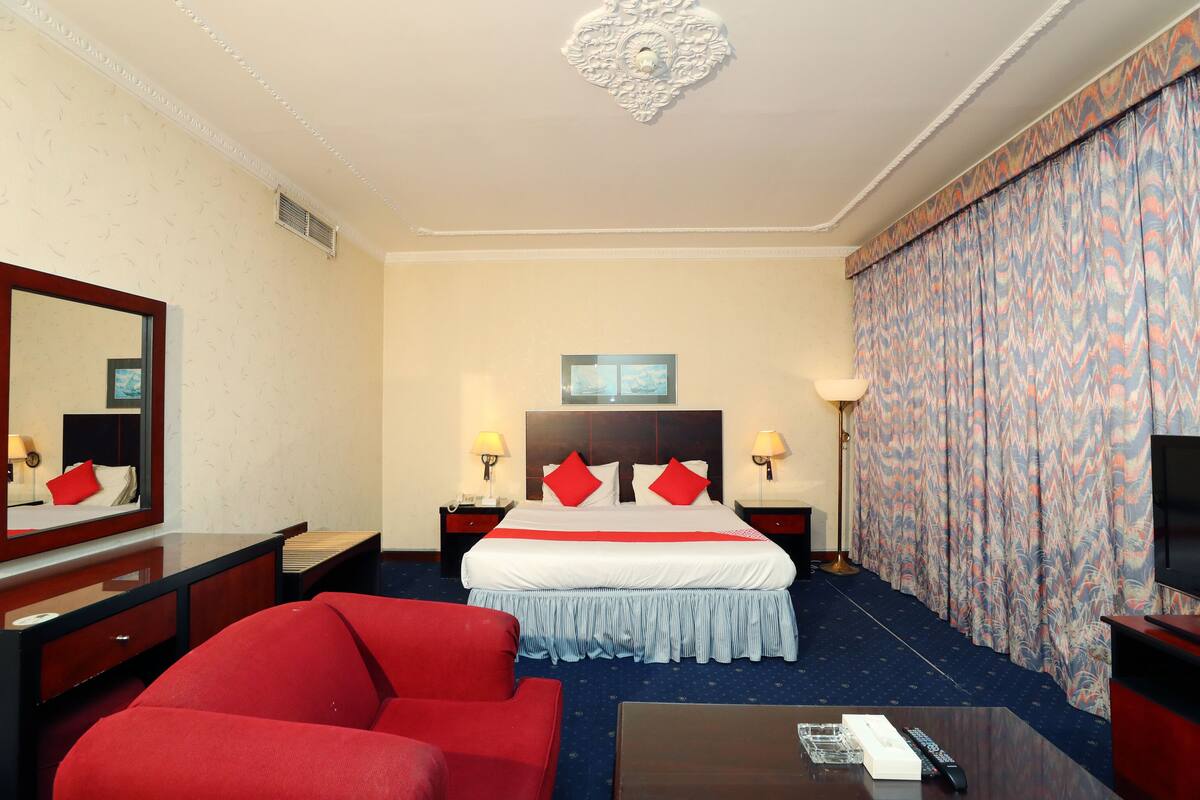 A hotel room with a large bed, two bedside tables with lamps, a TV, a red sofa, and a desk with a remote control.