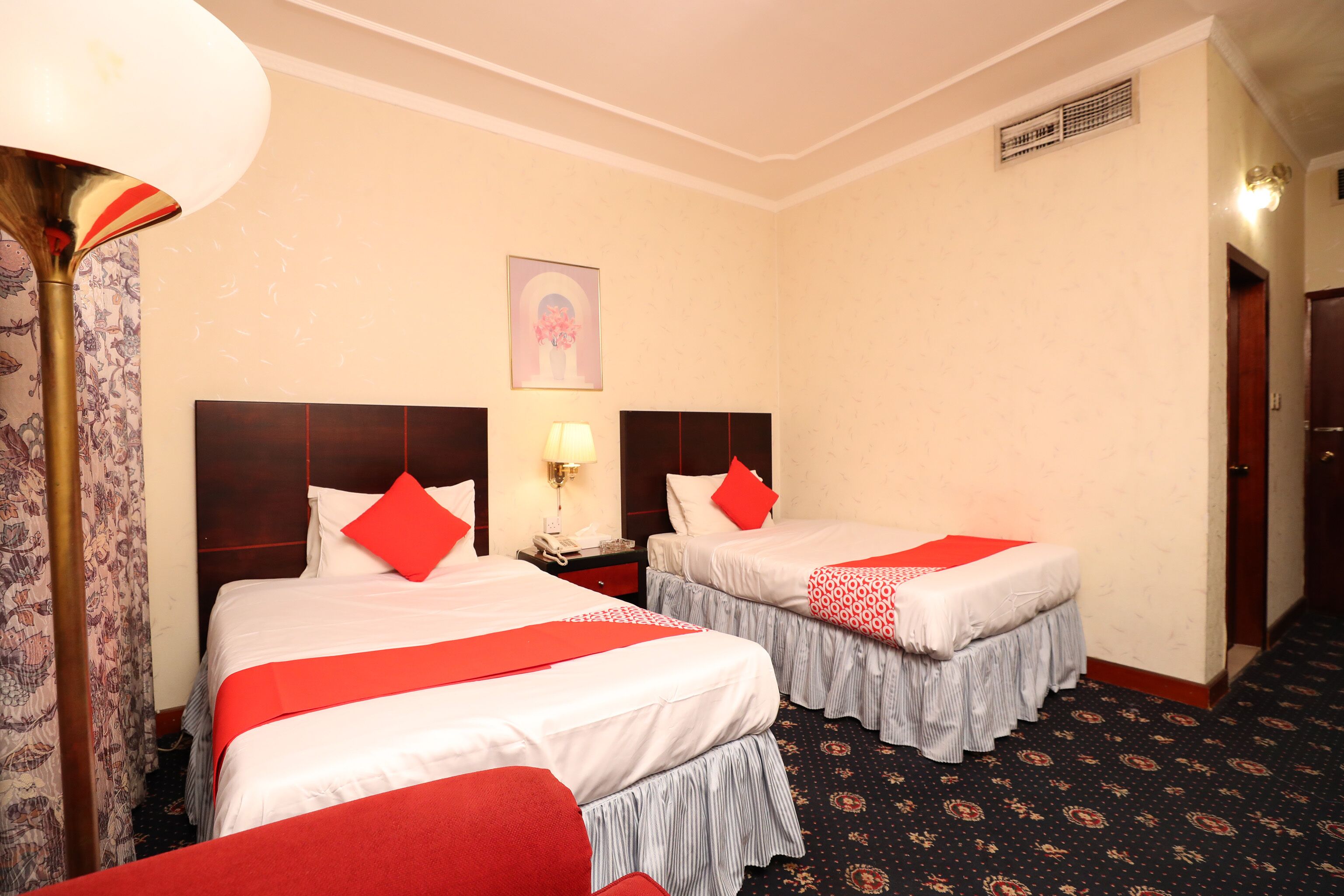A hotel room with two beds, each with red pillows and a white bedspread. There is a nightstand with a lamp and a phone between the beds. A painting hangs on the wall above the beds.