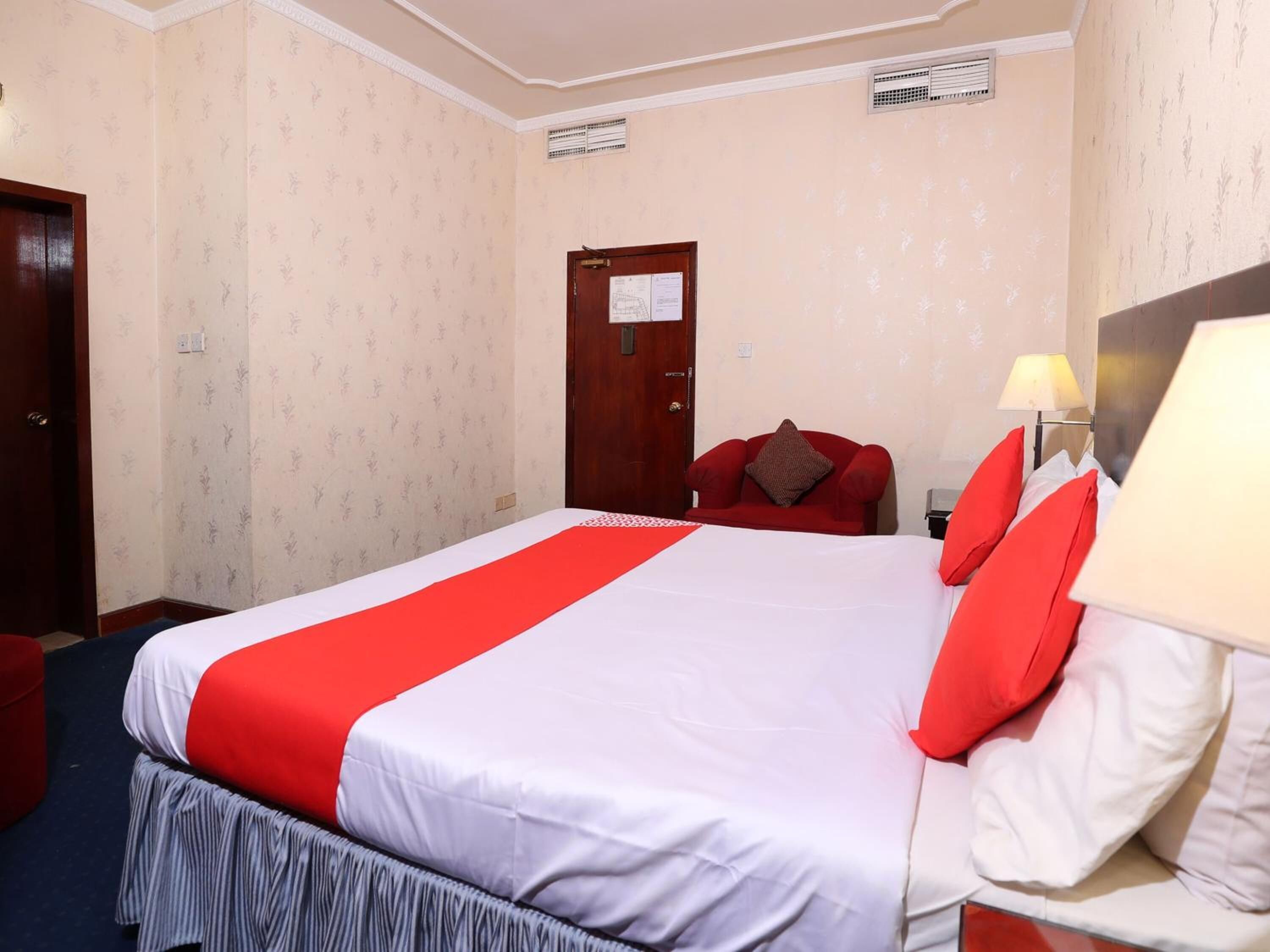 A hotel room with a large bed, red and white bedding, a red armchair, a desk with a lamp, and a door with a notice.