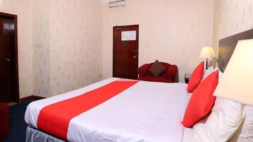 A hotel room with a large bed, red and white bedding, a red armchair, a desk with a lamp, and a door with a notice.