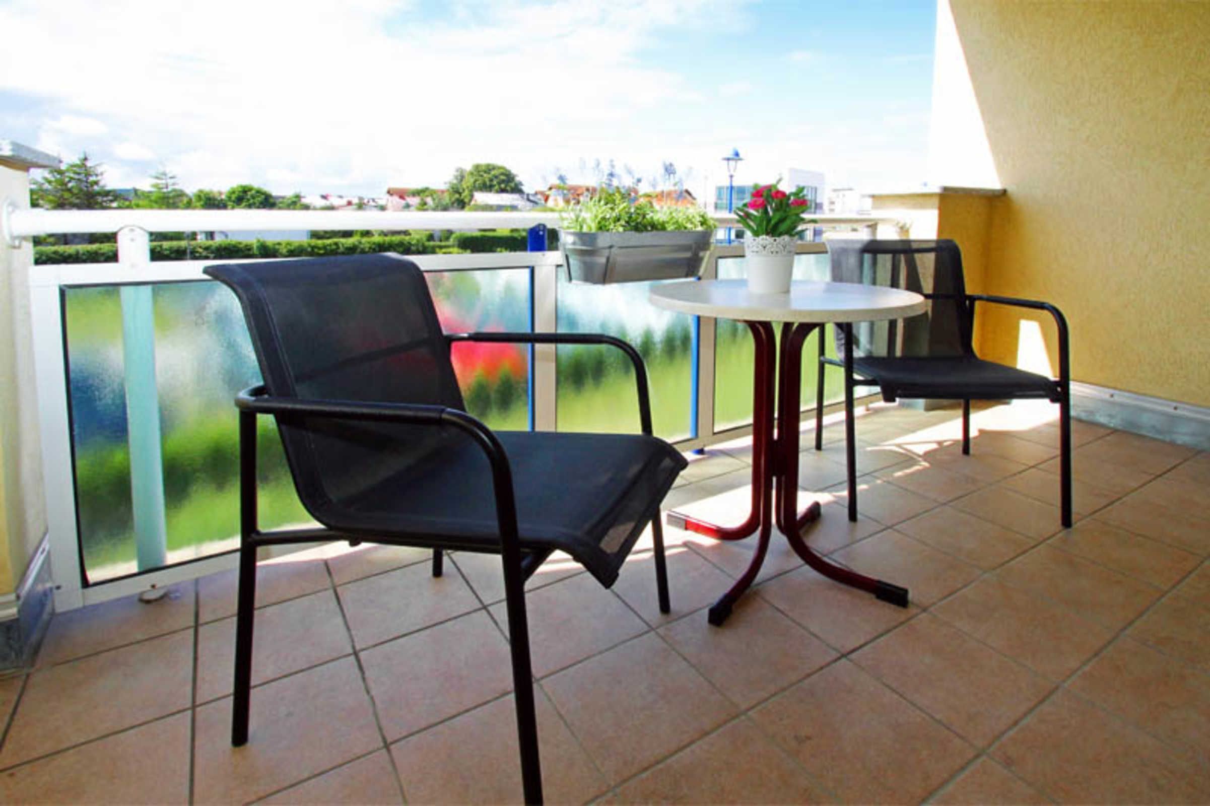 A balcony with two black chairs and a small round table, a potted plant, and a view of greenery and buildings.