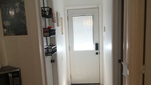 A hallway with a white door, a window with blinds, a microwave, a refrigerator, and a trash bin.