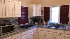 A kitchen with white cabinets, a granite countertop, a microwave, a stove, and a window with red shutters.