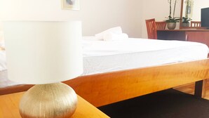 A hotel room with a bed, a wooden nightstand with a lamp, a black rug, a desk with a chair, and a potted plant.