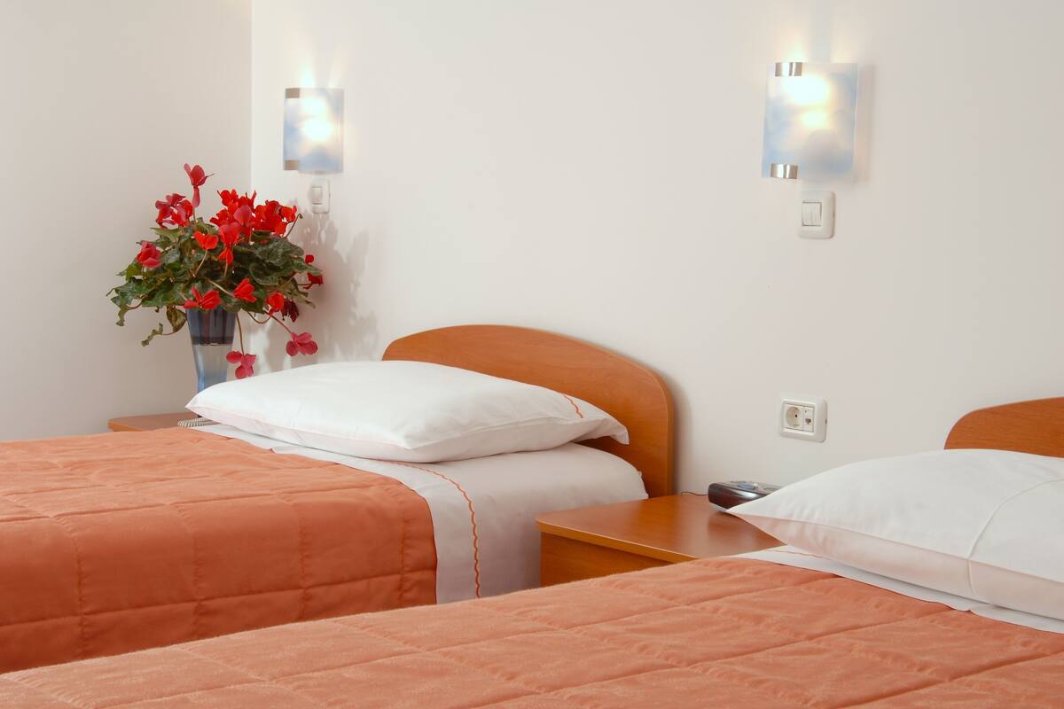 A hotel room with two beds, each with an orange bedspread, a white pillow, and a small bedside table. There is a vase with red flowers on one nightstand.