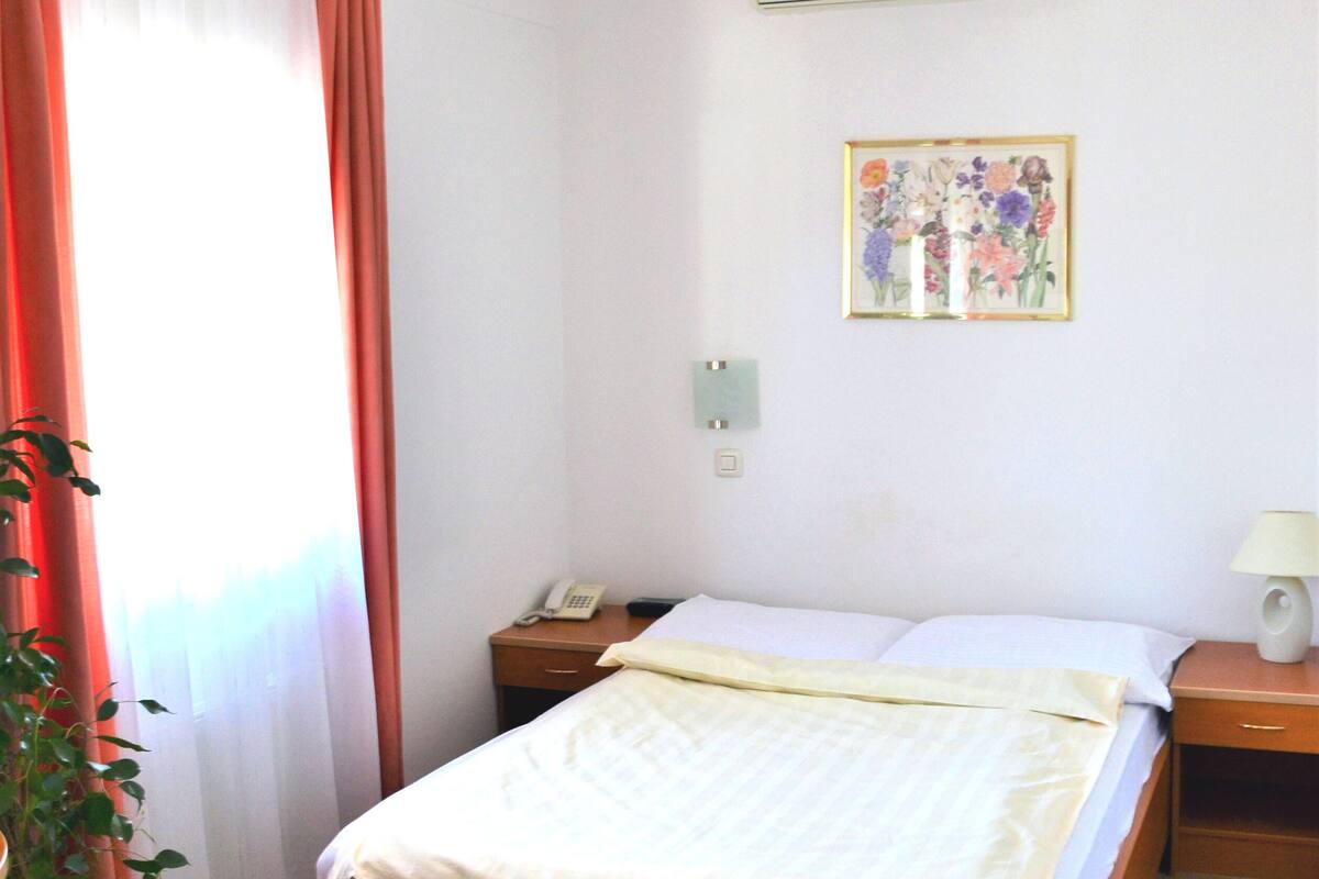 A hotel room with a wooden bed, a black mat, a potted plant, a painting, and an air conditioning unit.