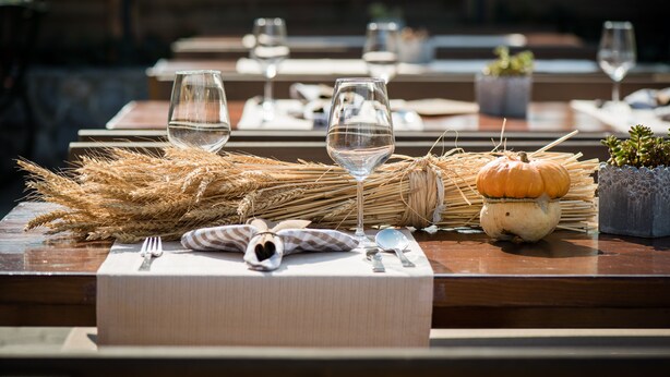 A dining table set with wine glasses, a small pumpkin, and wheat stalks.