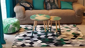Uma sala de estar com um sofá bege, um tapete estampado, uma mesa redonda pequena e uma luminária fixada na parede.