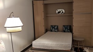 Uma cama compacta com colcha estampada, uma mesinha de cabeceira e uma luminária.