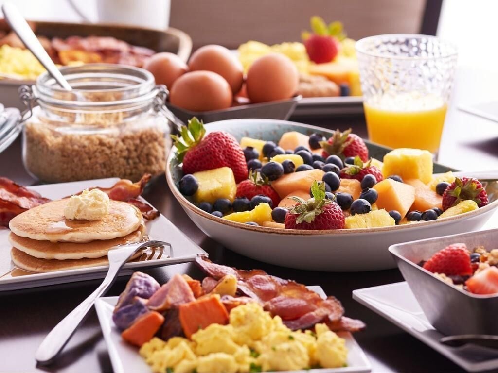 A breakfast spread with pancakes, scrambled eggs, bacon, and a variety of fruits and vegetables.