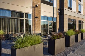 Un edificio moderno con ventanas amplias, mesas al aire libre y macetas con plantas verdes.
