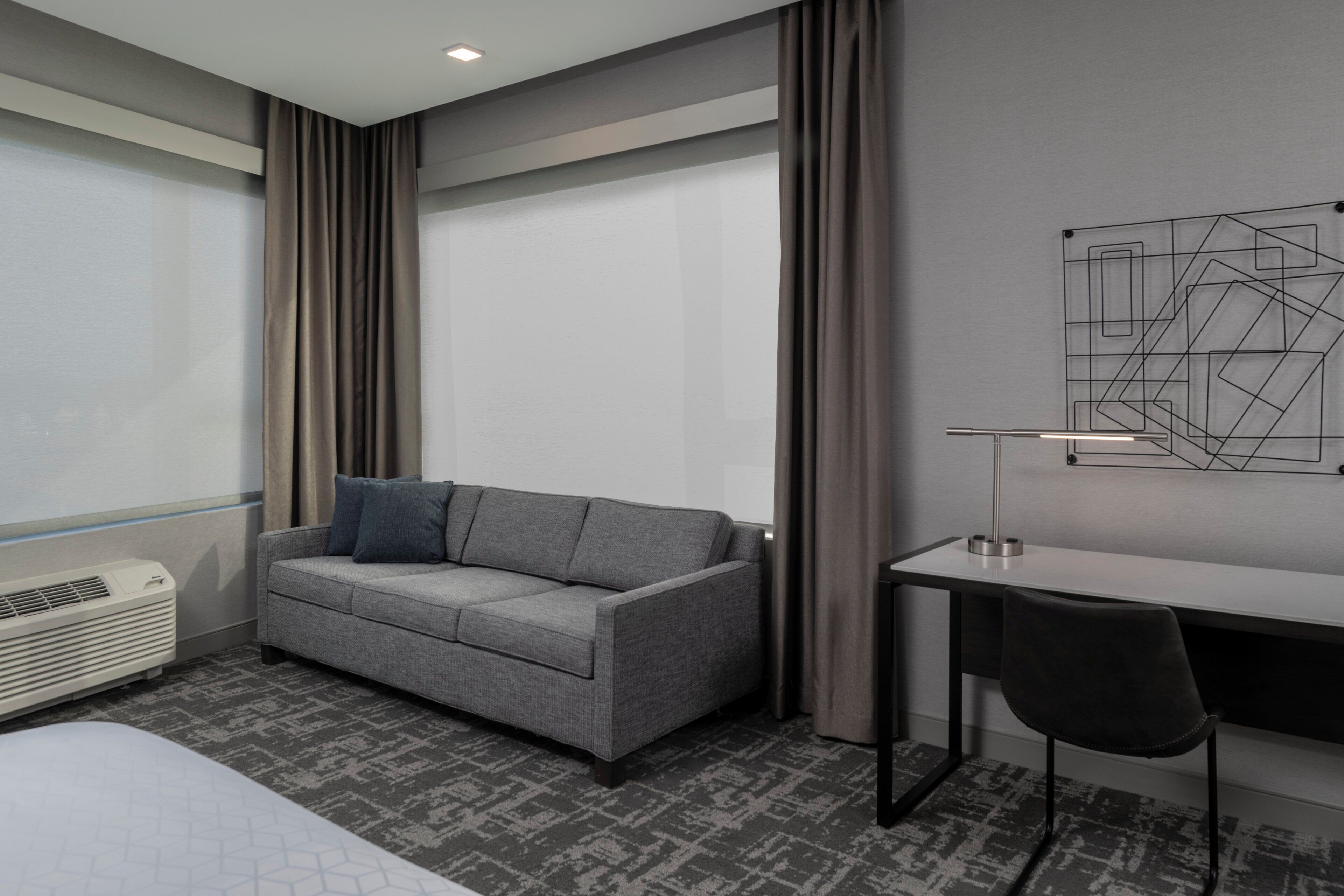 A modern hotel room with a grey sofa, a desk, a chair, and a large window with curtains.