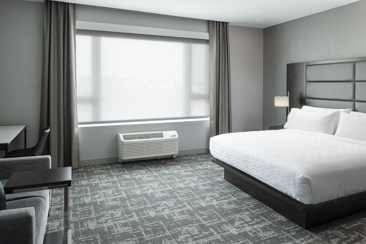 Une chambre d’hôtel avec un grand lit, un bureau, une chaise, une fenêtre avec des rideaux et une lampe fixée au mur.