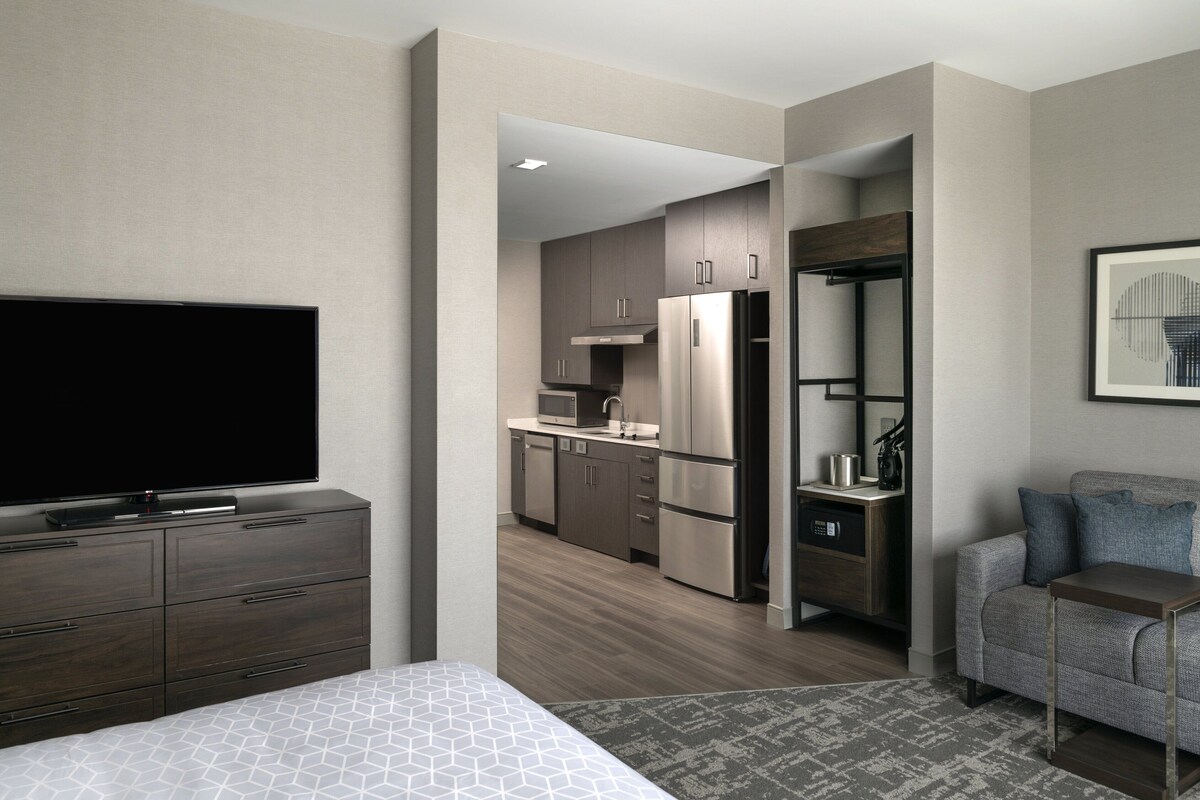 Une chambre d’hôtel moderne dotée d’un grand lit, d’une télévision à écran plat, d’une kitchenette et d’un coin salon avec un canapé et une petite table.