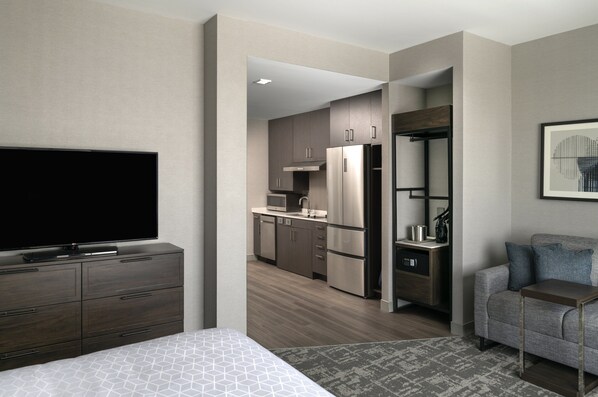 Habitación de hotel moderna con una cama grande, televisor de pantalla plana, kitchenette y zona de estar con sofá y una mesita.