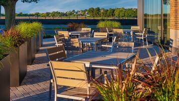 An outdoor patio with tables and chairs, surrounded by planters and a view of a body of water.